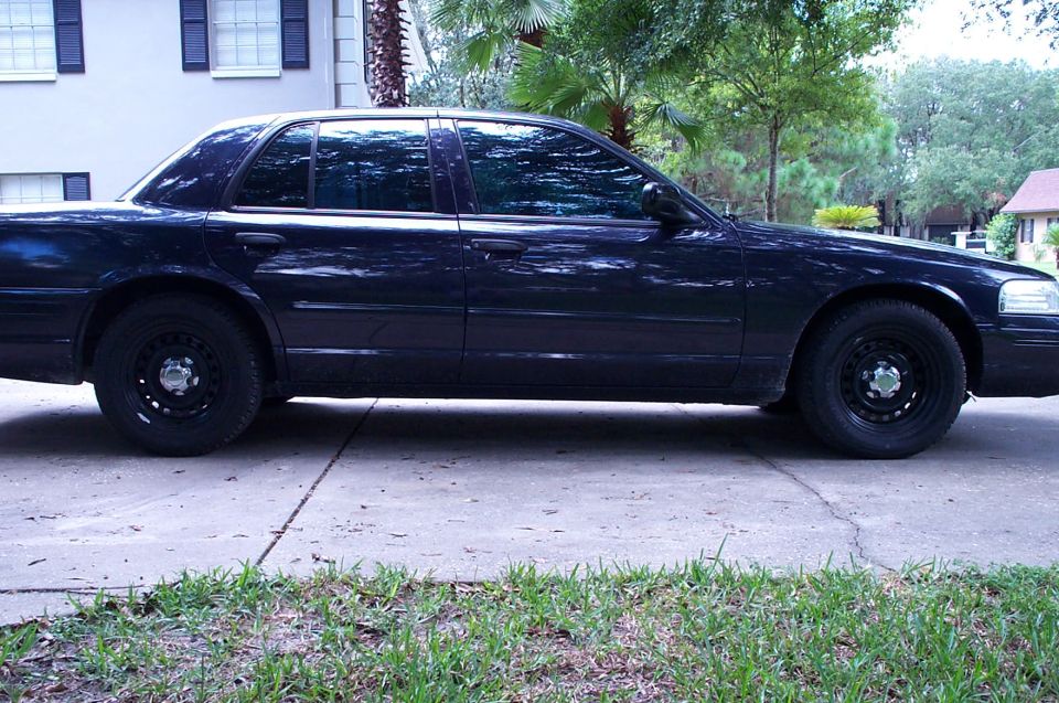Dark Ford Crown Victoria side profile on black steel wheels in a palm-lined driveway