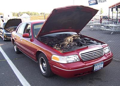 1999 Ford Crown Victoria - Two cars with hoods open parked side-by-side, one is a red Ford Crown Victoria.