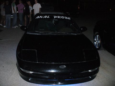 1991 Ford Probe - A black car with pop-up headlights is parked at night, with a banner on the windshield reading "ANAL PROBE".