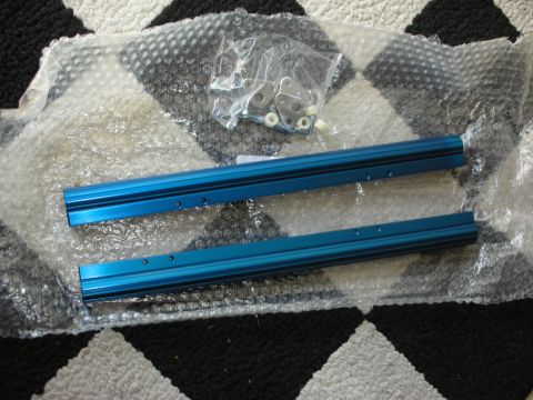 1998 Ford Crown Victoria - pair of blue anodized aluminum rails with drilled holes rest on bubble wrap next to a bag of mounting hardware on a checkered rug.