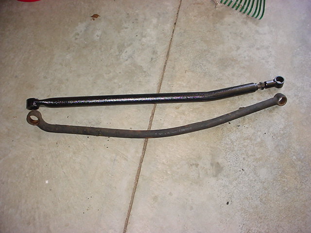 Two steering linkage bars, one black and one rusty, laid on a concrete floor
