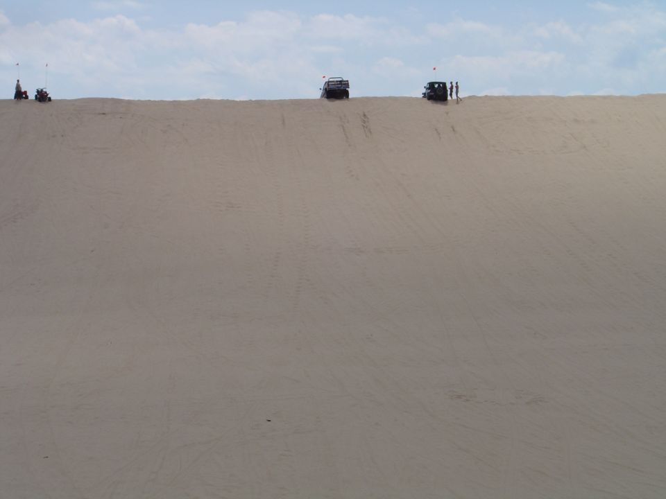 Several off-road vehicles and people on top of a large sand dune under a cloudy sky