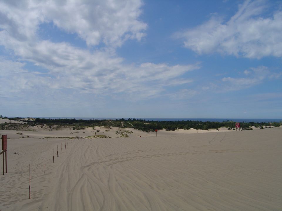 Sand dunes with a line of posts leading towards the ocean under a cloudy blue sky