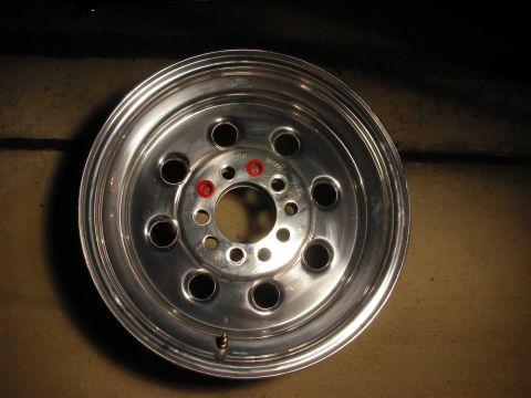 1998 Ford Crown Victoria - polished aluminum racing wheel with a multi-hole face and red lug-hole accents stands against a wall in dim lighting.