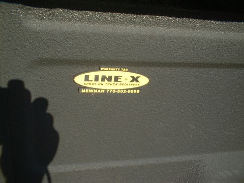 1995 Ford F-150 - Close-up of a textured gray spray-in bedliner inside a pickup bed, centered on the yellow oval LINE-X 'Spray-On Truck Bedliners' warranty t…