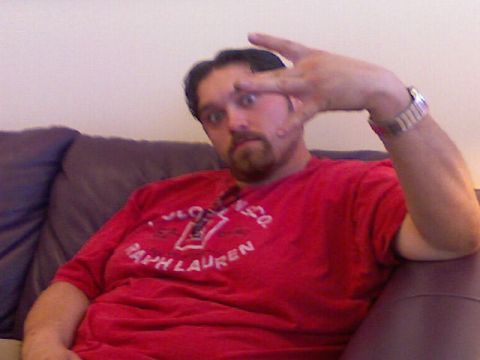 1996 Ford Probe - A man with a beard and goatee sits on a couch wearing a red t-shirt and a watch, making a peace sign with his hand.