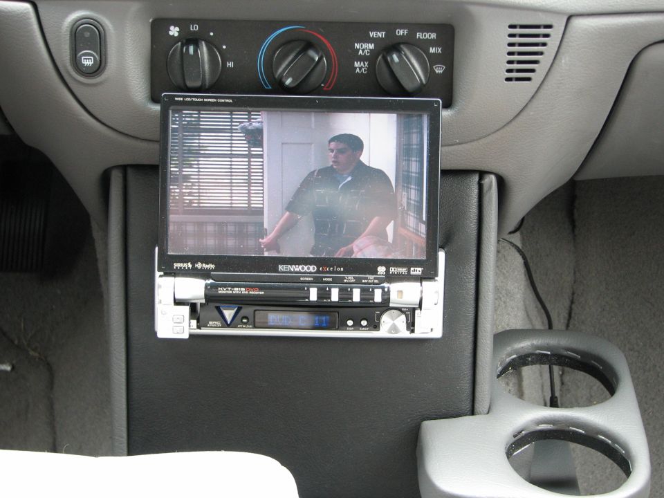 Car interior showing a Kenwood screen displaying a person, mounted above a car stereo, with cup holders to the right