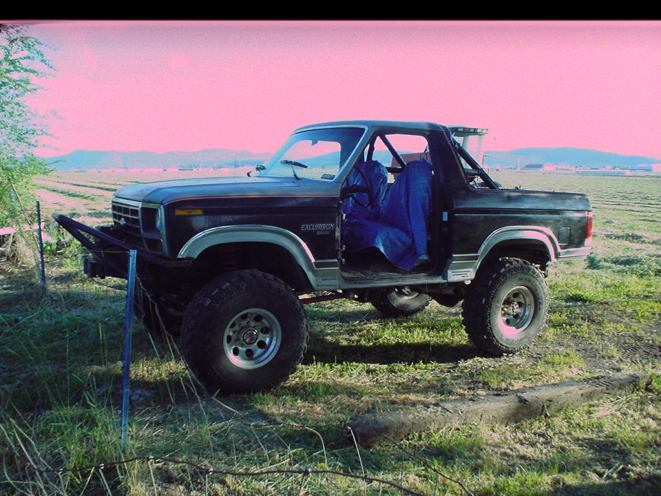 Lifted black Ford Bronco with no top and roll bar, on mud tires in a green field, photo tinted pink