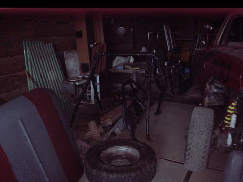 1984 Ford Bronco - A roll cage and spare tire are visible in a dimly lit garage, with parts of a red vehicle in the background.