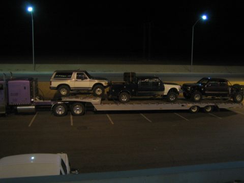 1990 Ford Bronco - purple semi truck and flatbed trailer loaded with a white SUV and two darker lifted trucks, parked at night.