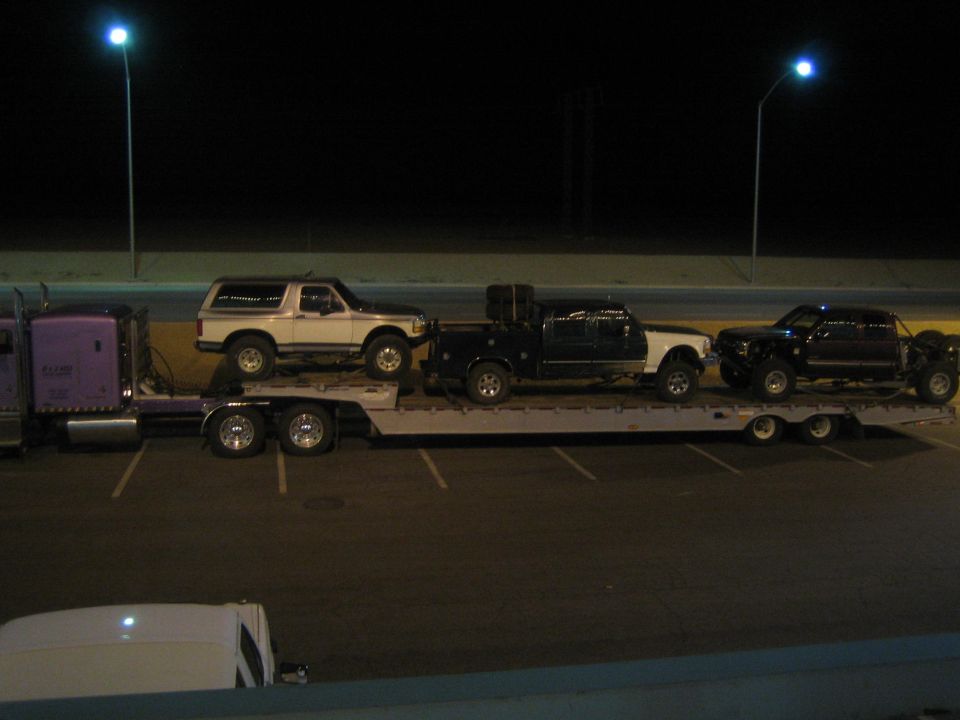 Purple semi with flatbed carrying white SUV and dark trucks at night