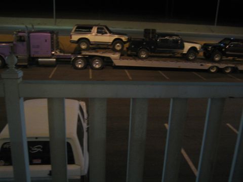1990 Ford Bronco - View through a white railing of a purple semi and flatbed trailer hauling a white SUV and trucks at night.