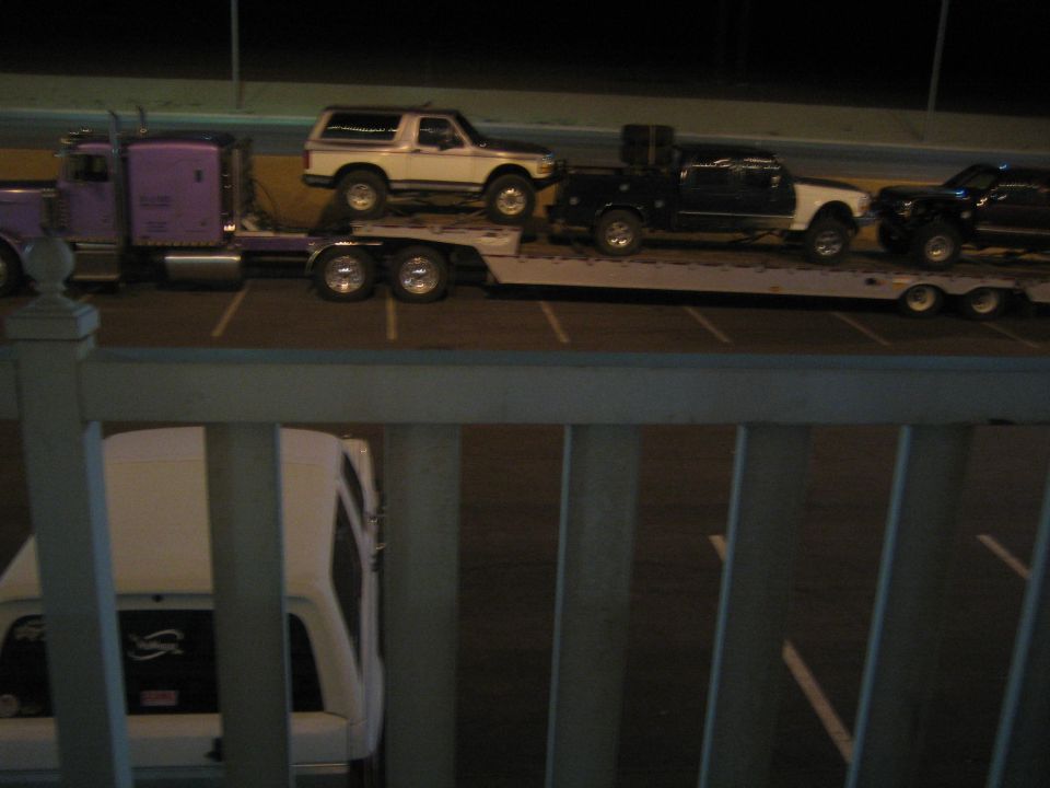 Purple semi hauling vehicles on flatbed seen through white railing at night