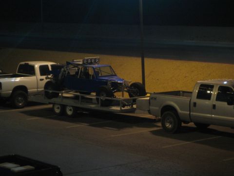 1990 Ford Bronco - blue tube-chassis off-road race buggy strapped to a trailer between two pickup trucks in a lot at night.