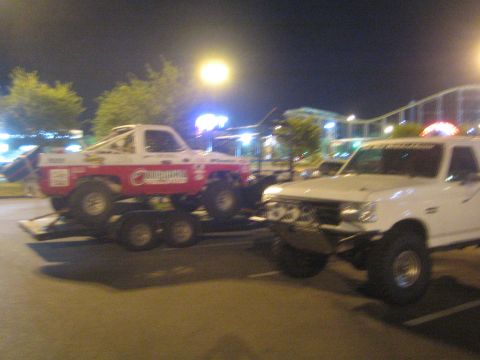 1990 Ford Bronco - white lifted pickup parked beside a red-and-white off-road race truck loaded on a trailer in a lit lot at night.