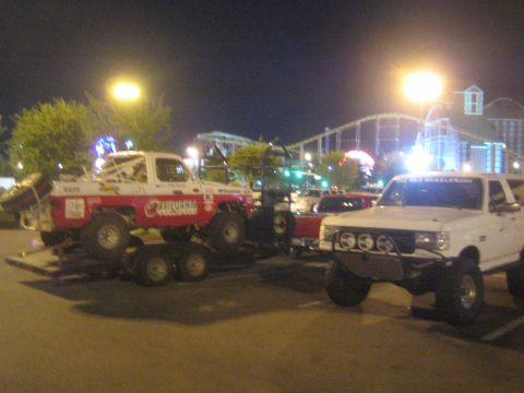 1990 Ford Bronco - white lifted pickup beside a red-and-white 'California' off-road race truck on a trailer, with amusement rides behind.