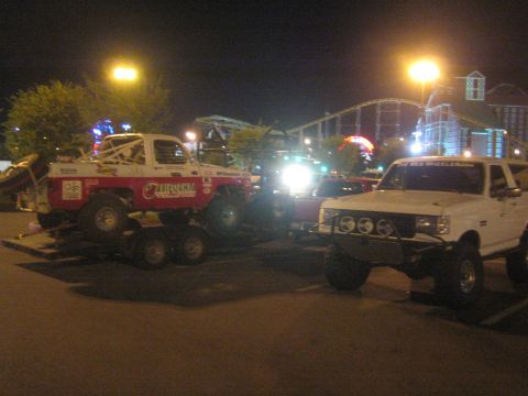 1990 Ford Bronco - white lifted pickup and a red-and-white race truck on a trailer at night, a rollercoaster lit up in the background.