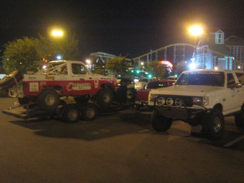 1990 Ford Bronco - white lifted pickup parked next to a red-and-white 'California' race truck loaded on a trailer in a lot at night.