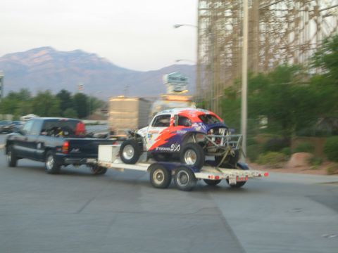1990 Ford Bronco - dark pickup towing a trailer carrying a blue-and-orange No. 990 race car along a road with mountains at dusk.