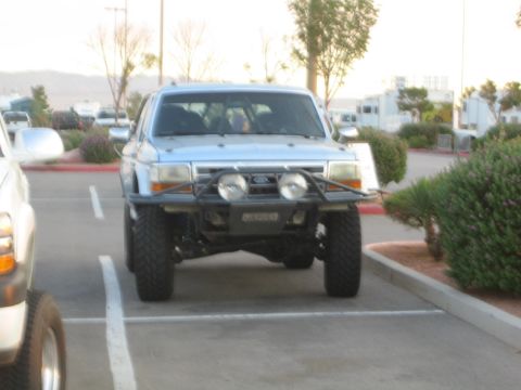 1990 Ford Bronco - silver lifted Ford pickup with a front brush guard and round off-road lights, parked in a sunny lot.