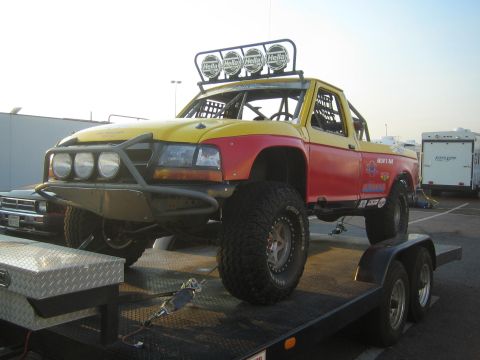 1990 Ford Bronco - yellow-and-red off-road prerunner truck fitted with a roof-mounted Hella light bar sits chained on a flatbed trailer.