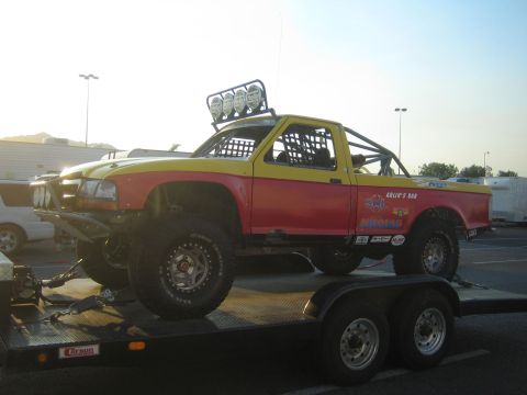 1990 Ford Bronco - Side view of a red-and-yellow off-road prerunner truck with a roll cage and roof light bar strapped onto a tandem-axle trailer.