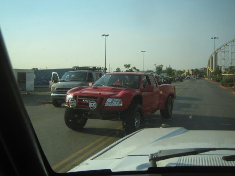 1990 Ford Bronco - red lifted prerunner pickup with bumper-mounted light pods crosses a road, with a roller coaster visible in the distance.