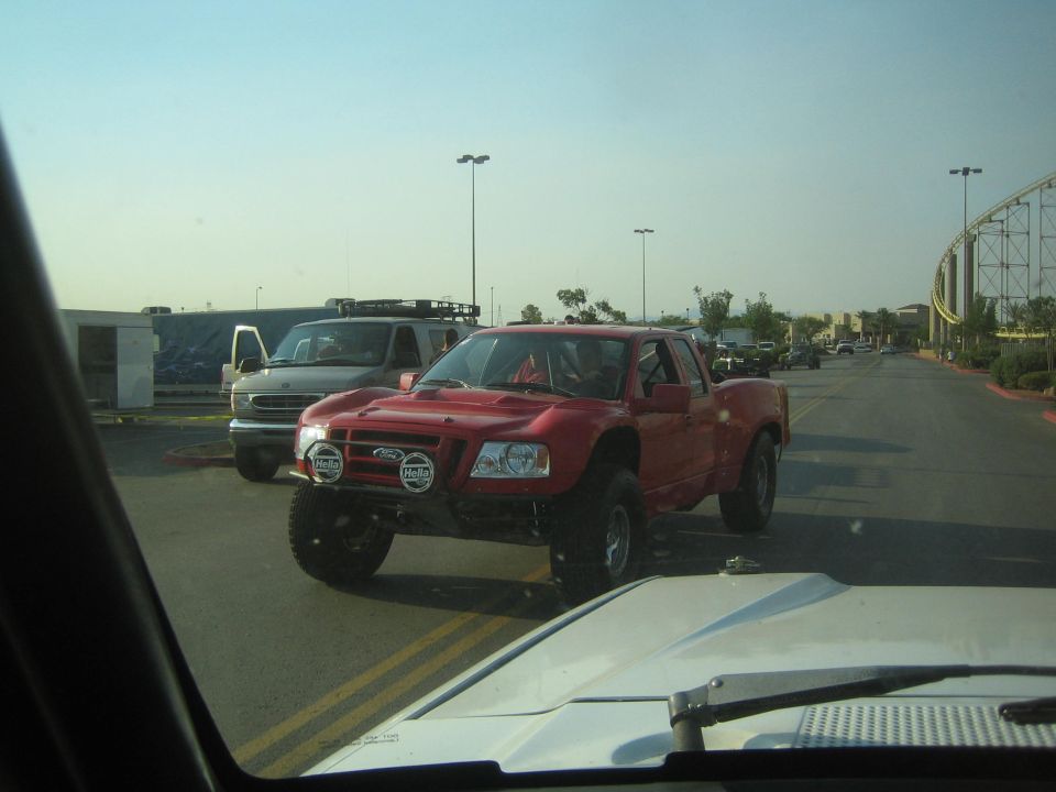 Red lifted prerunner pickup with light pods crossing a road near a roller coaster