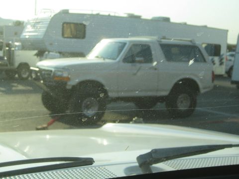 1990 Ford Bronco - white lifted Ford Bronco sits in a paddock lot beside RVs, viewed through a dirty windshield with a wiper visible.