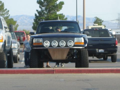 1990 Ford Bronco - dark blue lifted prerunner truck with four front Hella light pods sits in a lot as people stand nearby.