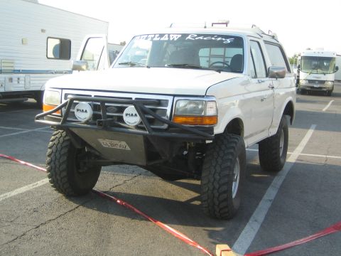 1990 Ford Bronco - white lifted Ford Bronco fitted with PIAA driving lights and an Outlaw Racing windshield banner sits in a desert paddock.