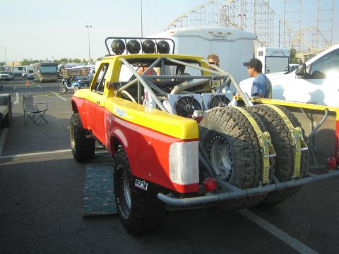 1990 Ford Bronco - Rear view of a yellow-and-red prerunner truck carrying twin spare tires on a rear rack with a roof light bar, parked at an event.