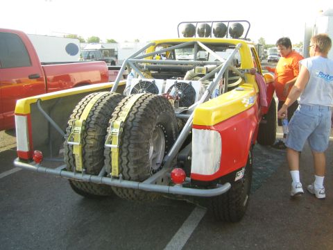 1990 Ford Bronco - Rear of a yellow-and-red tube-frame prerunner truck with dual rear spare tires while two men stand beside it in a paddock.