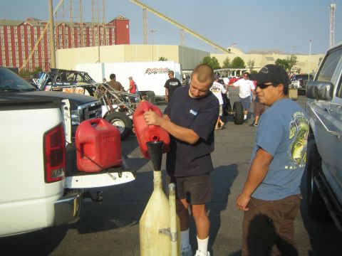 1990 Ford Bronco - man pours fuel from a red gas can through a long funnel at a desert race paddock as another man watches.