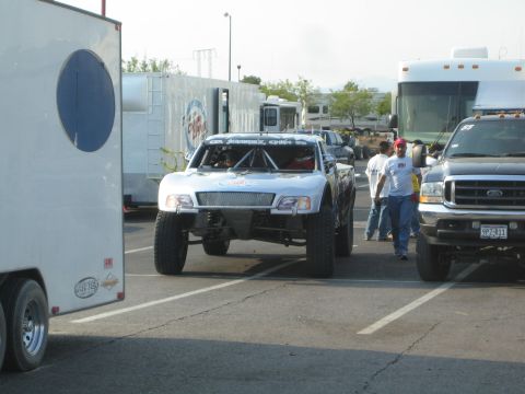 1990 Ford Bronco - white off-road prerunner truck drives through a crowded paddock between an enclosed trailer and a bus.