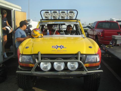 1990 Ford Bronco - Front view of a yellow-and-red prerunner truck with a four-light Hella roof bar and SHL hood logo as crew gather around.