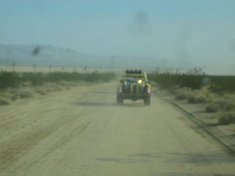 1990 Ford Bronco - yellow off-road race vehicle speeds away down a dusty desert dirt road seen through a hazy windshield.