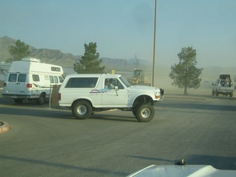 1990 Ford Bronco - white lifted Ford Bronco drives through a dusty desert lot near a camper van and RVs.