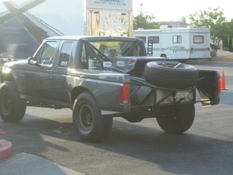 1990 Ford Bronco - Rear three-quarter view of a gray Ford-based prerunner truck with a bed-mounted spare tire and tube bumper at a Reno off-road event.