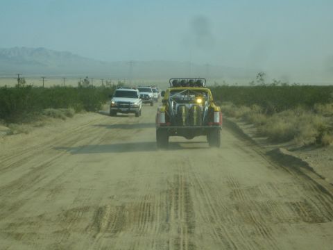 1990 Ford Bronco - yellow open buggy and a white SUV travel down a dusty desert dirt road with mountains on the horizon.