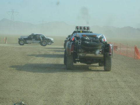 1990 Ford Bronco - Two off-road race trucks, one numbered 58, run on a dusty desert course lined with orange safety fencing.