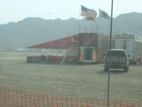 1990 Ford Bronco - red-and-white grandstand structure flying American flags stands across a dusty desert race venue behind orange fencing.