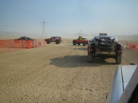 1990 Ford Bronco - Several off-road race trucks are staged on a dusty desert course beneath power lines, lined by orange safety fencing.