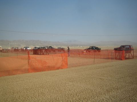 1990 Ford Bronco - Several off-road trucks and buggies staged behind orange safety fencing on a flat desert pit area with distant mountains.