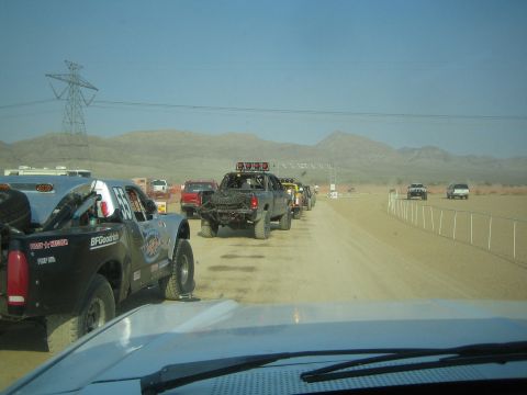 1990 Ford Bronco - blue off-road race truck with spare tires follows other race trucks down a dusty desert road below power lines.