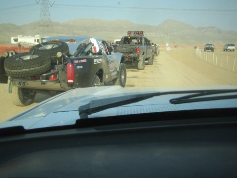 1990 Ford Bronco - black off-road race truck carrying two spare tires leads a line of vehicles on a dusty desert course.