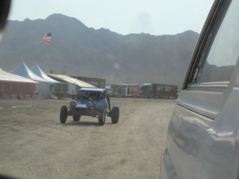 1990 Ford Bronco - blue open dune buggy crosses a dusty staging lot lined with vendor tents, trailers and a large American flag.