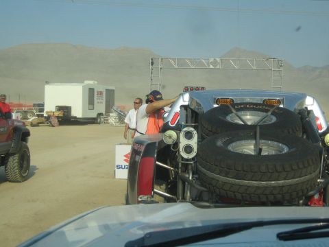 1990 Ford Bronco - rear of an off-road race truck with a mounted spare tire sits near officials and a Suzuki banner in a dusty pit.