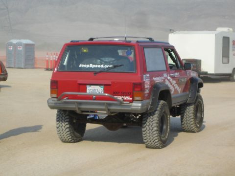 1990 Ford Bronco - red lifted two-door Jeep Cherokee with oversized tires and a JeepSpeed windshield banner parked on a dusty lot.