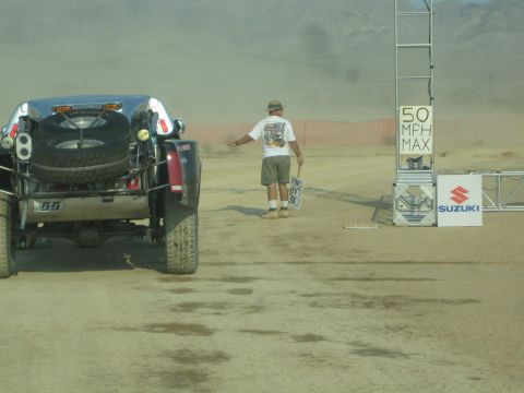 1990 Ford Bronco - off-road race truck waits at a start gate marked 50 MPH MAX while an official gestures beside a Suzuki banner.